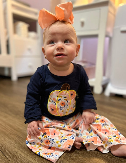 Girls' Navy Pumpkin Bell Bottom Set: A navy blue shirt with a large floral pumpkin design and ruffles on the sleeves, paired with bell-bottom style pants featuring a matching floral pattern.